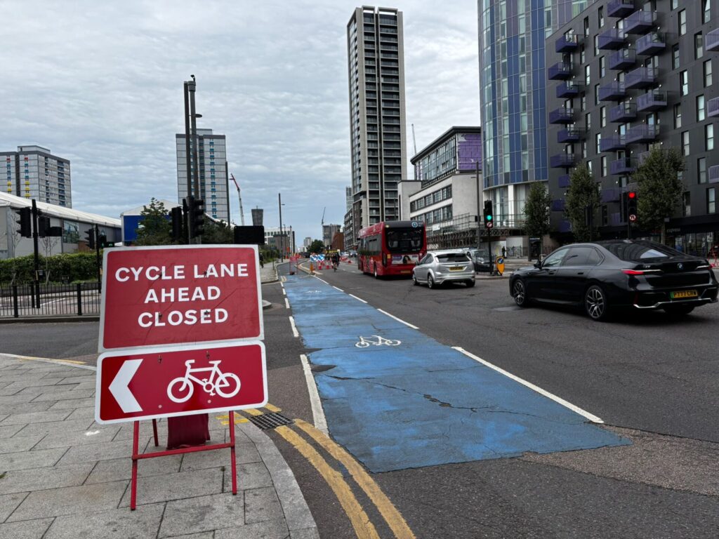 A cycle lane through a large junction with a sign saying "CYCLE LANE AHEAD CLOSED" and a diversion arrow pointing to the left. Ahead the cycle lane is closed with traffic cones directing cyclists into the traffic lane in front of a bus.