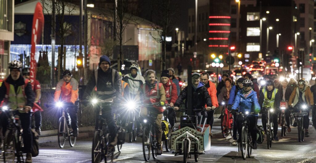 Lots of different people cycling in the evening on a major road, with lights twinkling, on a protest. The rider at the front has a London Cycling Campaign sail flag on the back of his bicycle.