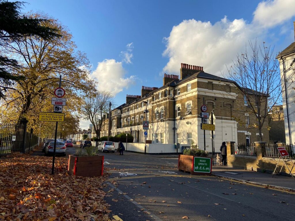 A residential street with autumn leaf-fall. A filtered road closure with "no motor vehicles except permit holders WHP01" signs, planters narrowing the road, camera enforcement, and a "road open to pedestrians, kick scooters, wheelchairs, bicycles" sign. Gorgeous autumn/early winter light shines through the trees.