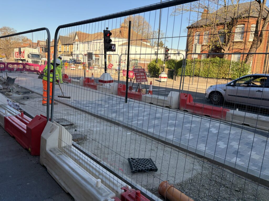Through metal fencing, workers sweep dust from the top of a new traffic island separating a one way cycle track from traffic.