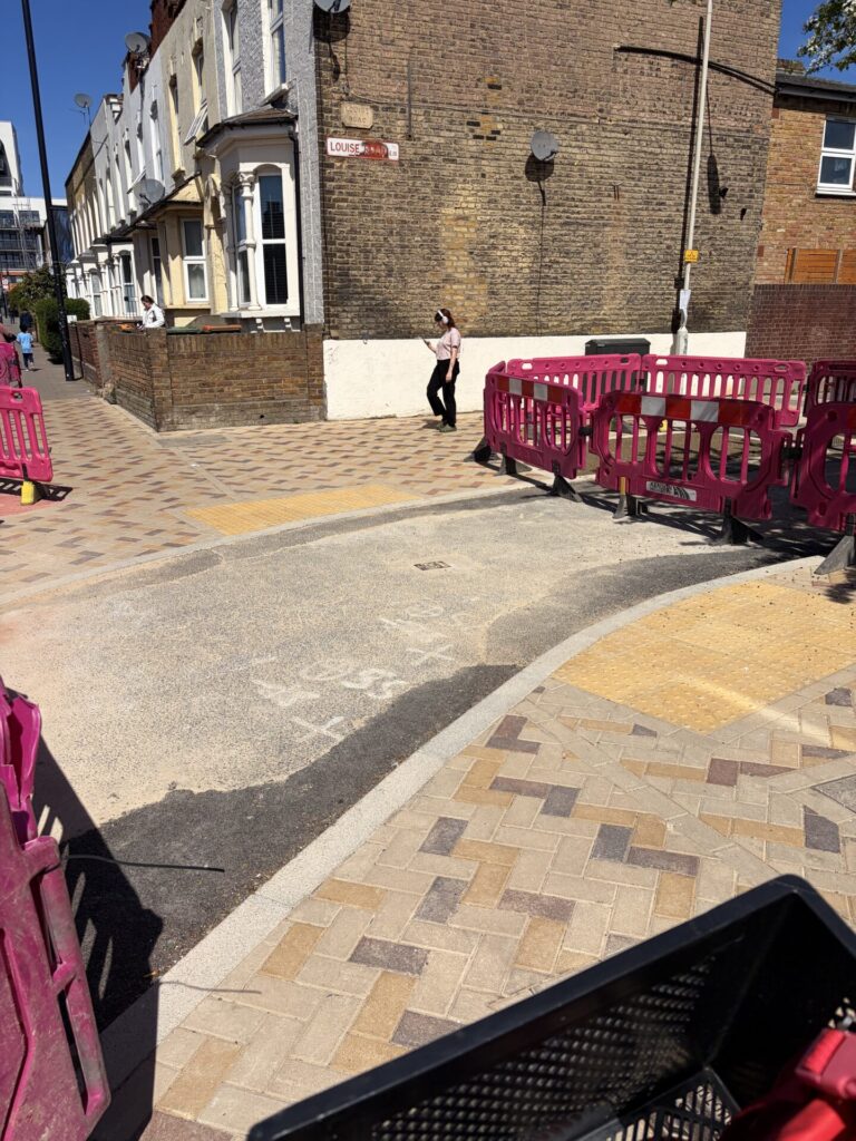 Curved cycle path through a pavement with a crossing marked with yellow rectangular tactiles. The pavement is in jazzy brown, gold and ochre-coloured block paving.