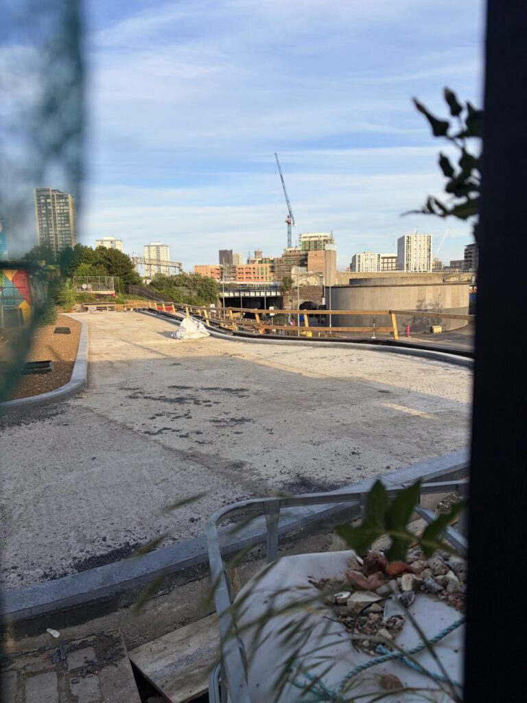 Taken through fencing: The kerbing for a wide switchback ramp with a sandy/concrete substrate. Wooden barriers prevent falls at the side.