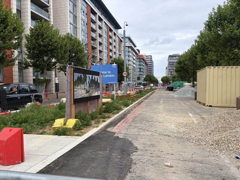Rain gardens in a median with a signboard showing a visualisation of the final scheme. The future cycle track retains the carriageway surfacing for now.