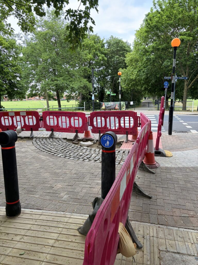 Footway with setts in an arc pattern being reconstructed behind plastic fencing.
