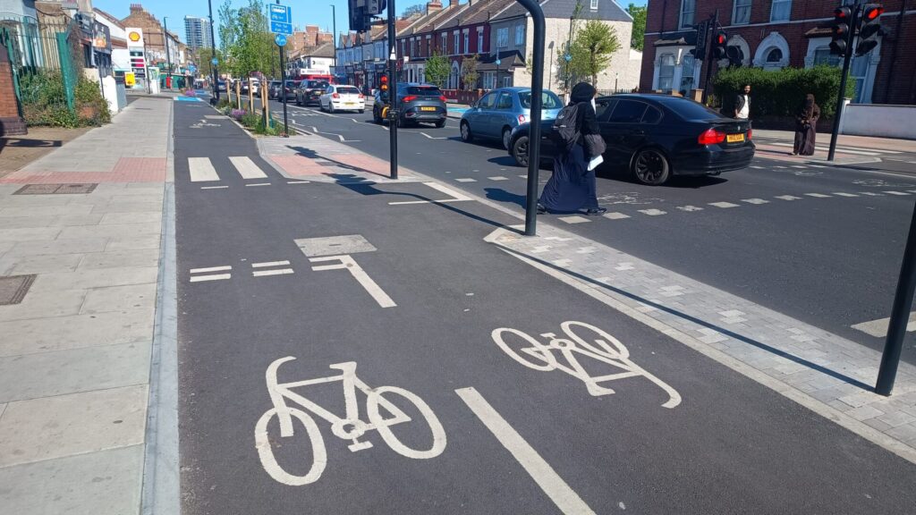 The one-way cycle track with a signalised crossing of a carriageway leading off to the right, with a short 2-way stub of cycle track in the foreground. The signalised crossing is a parallel signalised pedestrian and cycle crossing, accessible for pedestrians from a median using a zebra crossing of the cycle track. The zebra crossing is marked with red tactile in an L shape towards the back of the footway.
