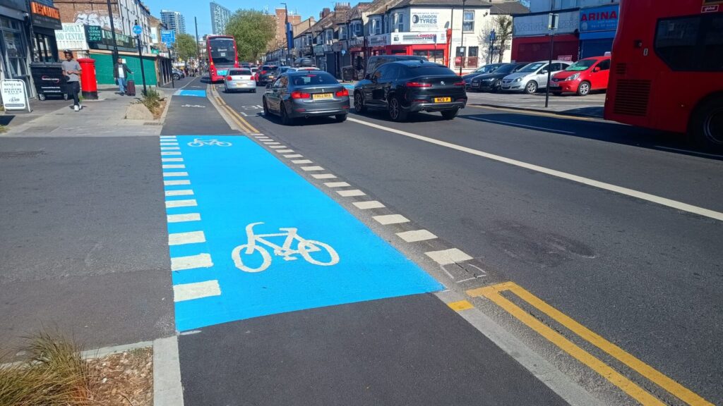 A one-way cycle track on a street with rain gardens and a vehicle crossover on the left (through which the cycleway is marked with blue paint), and a bus lane on the right.