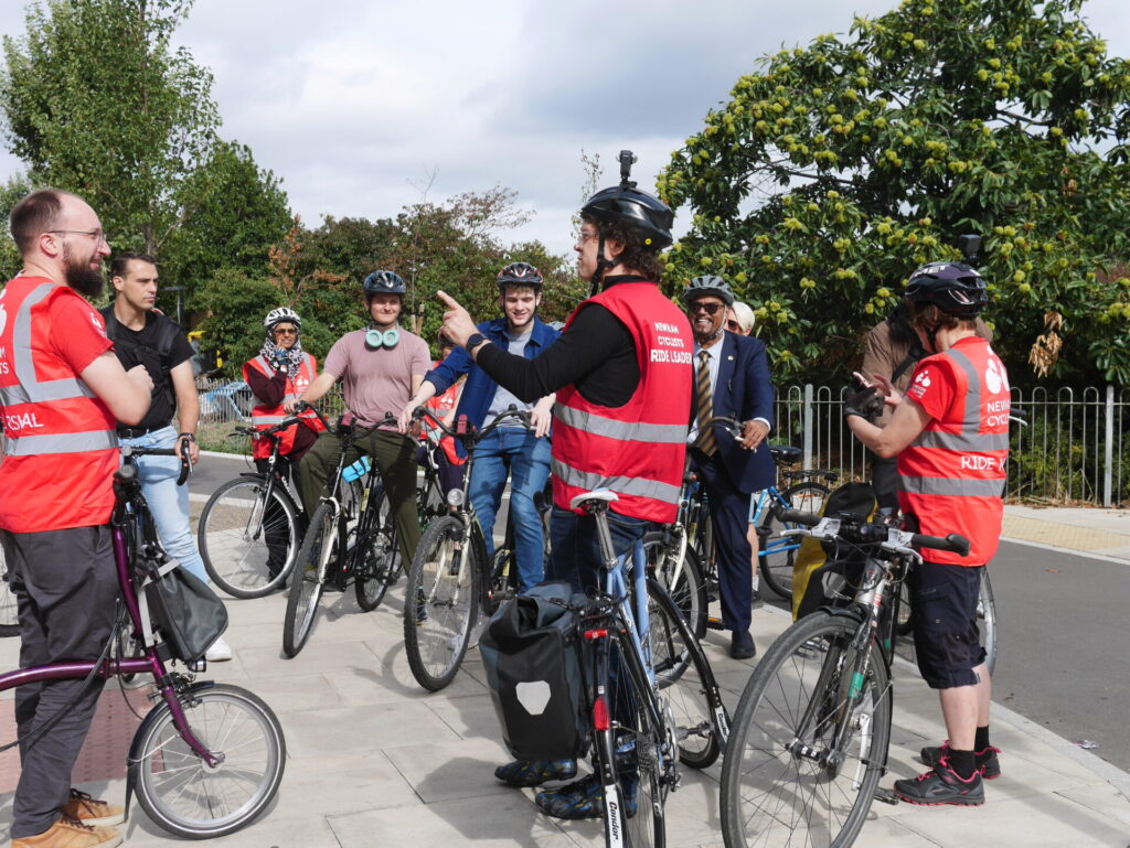A group of people on bicycles talking about a freshly-opened protected junction: Jonathan, Shameem, Richard, and Karen from Newham Cyclists; also Cllr John Morris, Cllr Ren Tilbury, Cllr Nate Higgins, Cllr Mehmood Mirza, and others in the background.
